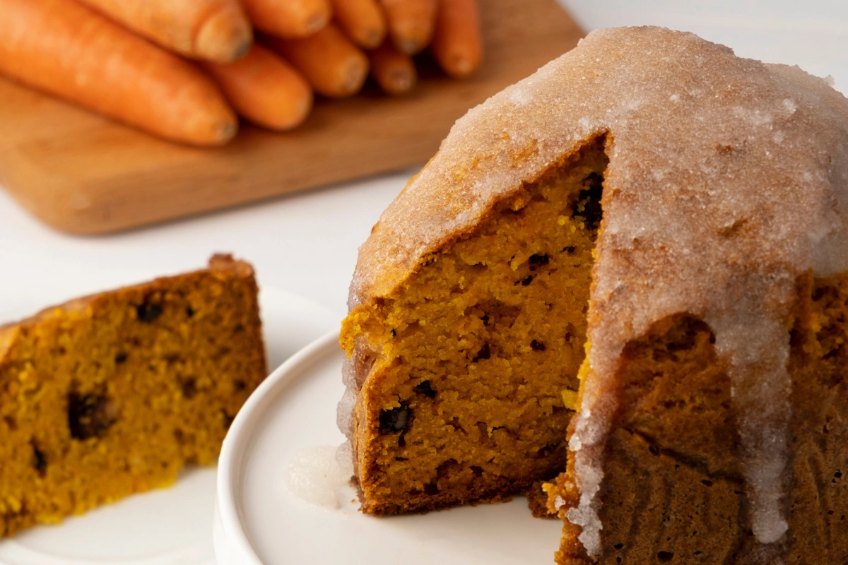 Moist vegan carrot cake with a slice cut out, showcasing its golden interior and topped with a light glaze, with fresh carrots in the background.