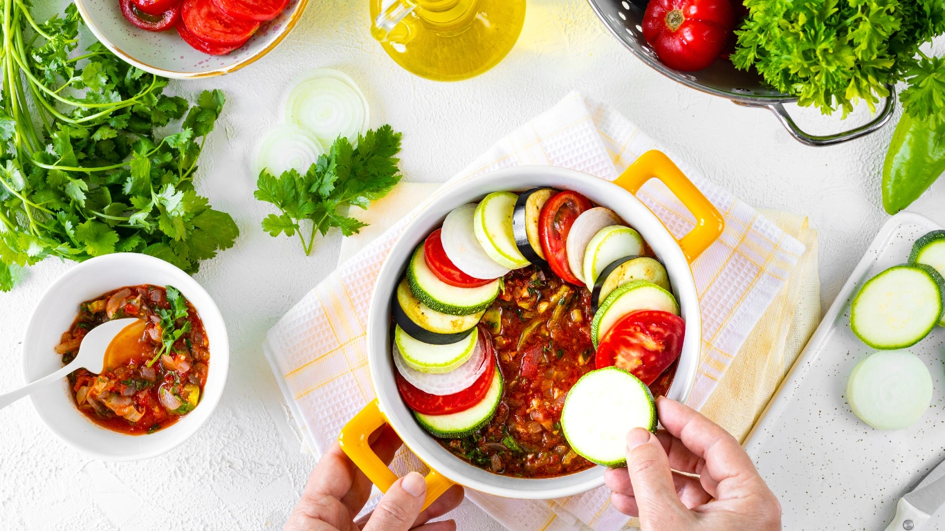 A vibrant vegan dish in a yellow baking dish with zucchini, tomatoes, and eggplant slices, surrounded by fresh herbs and ingredients on a white countertop.