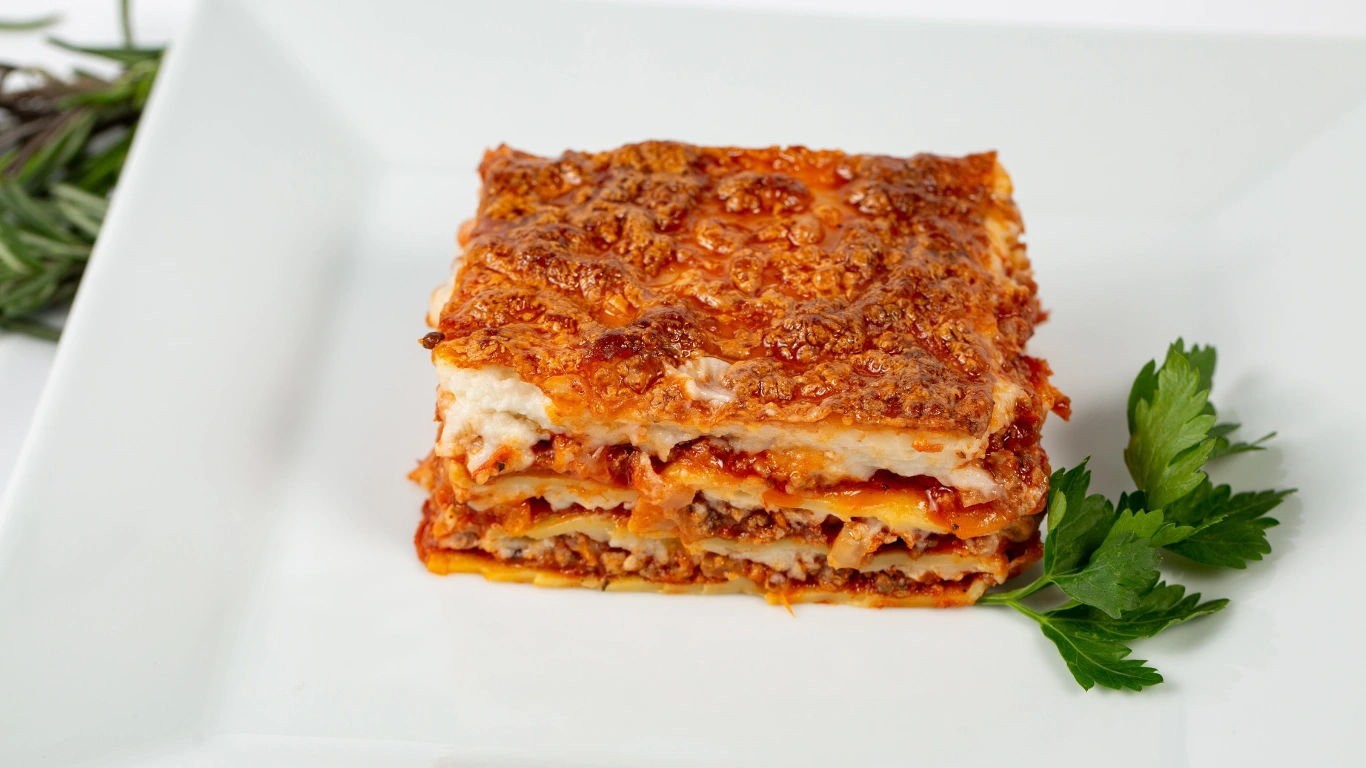 A beautifully plated slice of vegan lasagna with golden-brown layers of noodles, rich tomato sauce, and creamy topping, garnished with fresh parsley on a white plate.