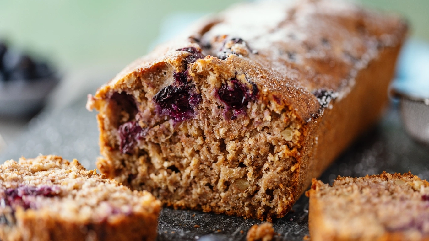 Close-up of a freshly baked vegan blueberry loaf cake with visible blueberries and a golden-brown crust, sliced for serving.