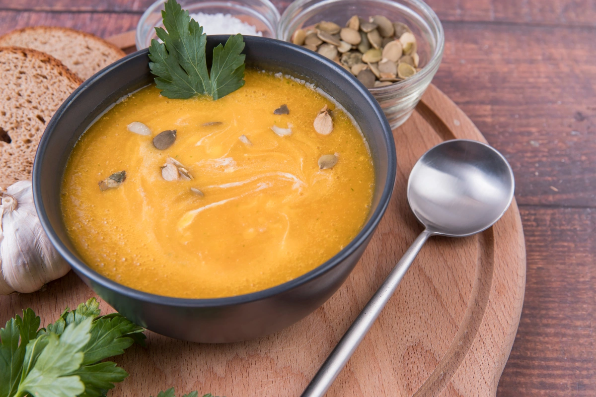 Peanut and pumpkin puree soup garnished with pumpkin seeds and parsley, served in a black bowl on a wooden plate with bread and garlic.