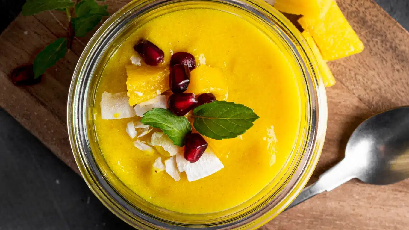 Bright yellow mango dessert garnished with pomegranate seeds, coconut flakes, and fresh mint leaves in a glass jar.
