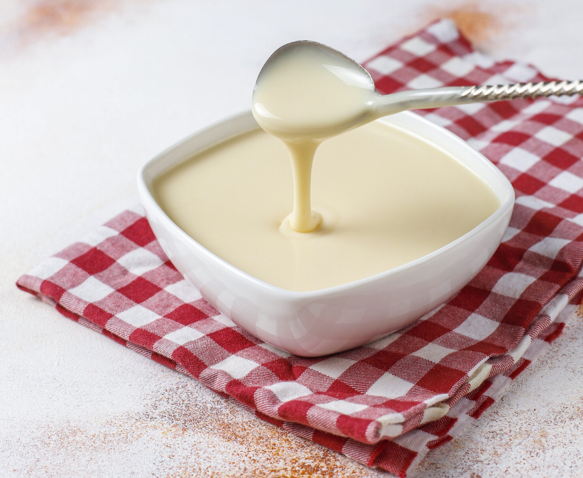 A spoon pouring creamy vegan cashew glaze into a white bowl, placed on a red and white checkered cloth on a light textured surface.