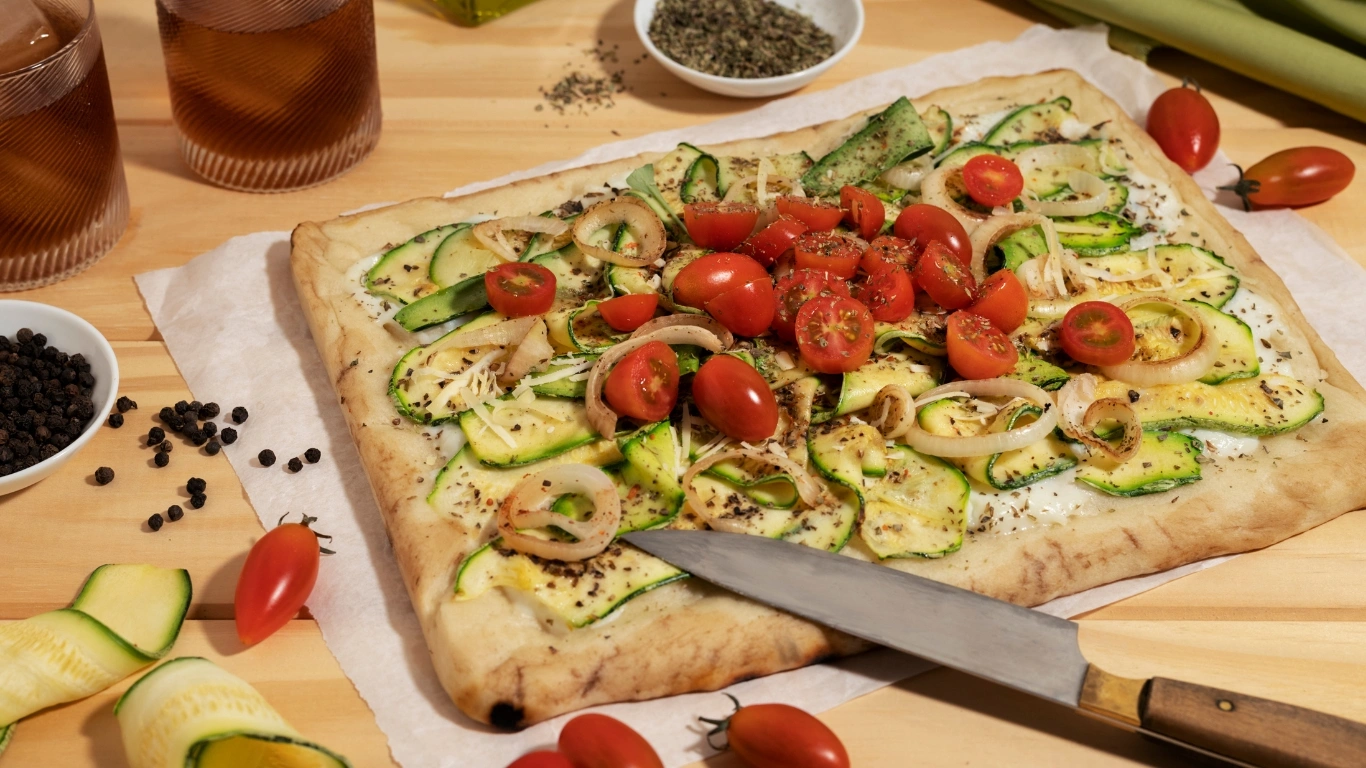 Vegan cauliflower pizza crust topped with fresh zucchini slices, cherry tomatoes, and onions, served on a parchment-lined wooden surface.
